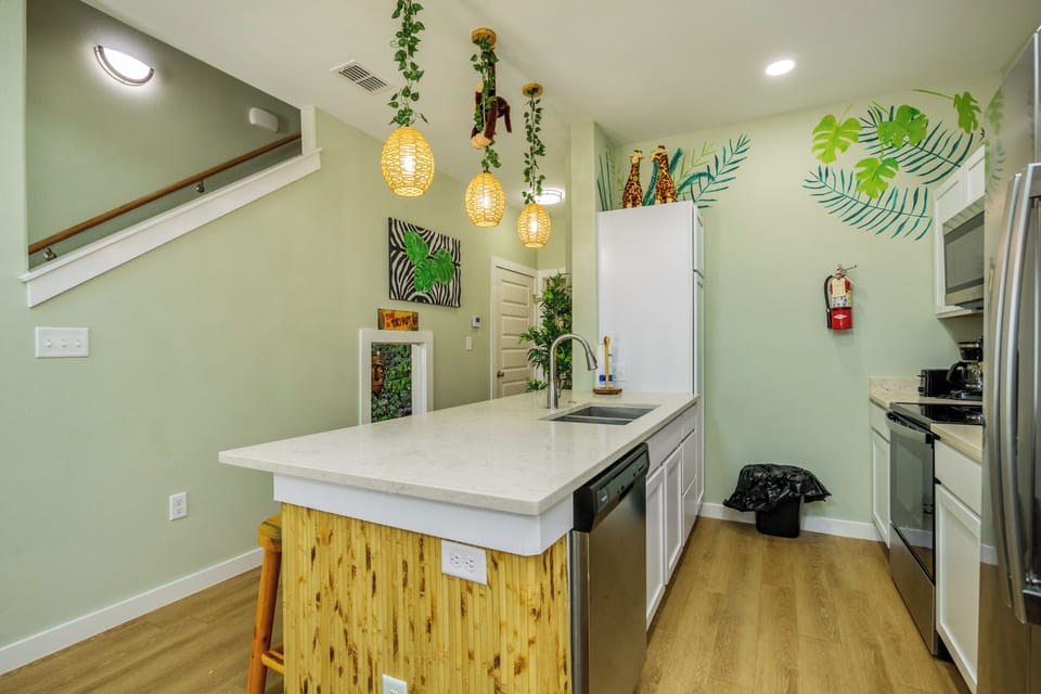Your tropical-themed kitchen invites culinary adventures with sage walls, bamboo accents, and modern appliances beneath charming pendant lights.