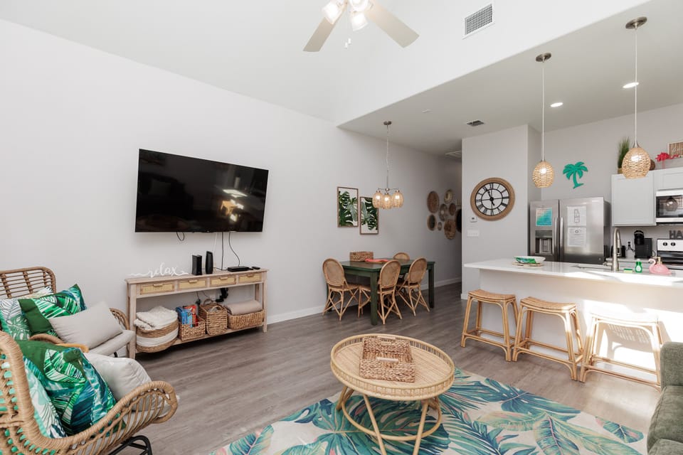 Relax in this bright tropical-themed living space featuring natural rattan furniture, palm leaf accents, and an open kitchen perfect for your vacation meals.
