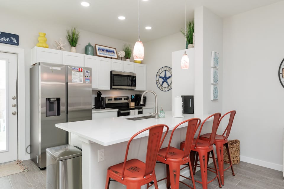 Gather around your stylish breakfast bar with vibrant coral stools while enjoying modern appliances and coastal-inspired décor that makes cooking together a joy.