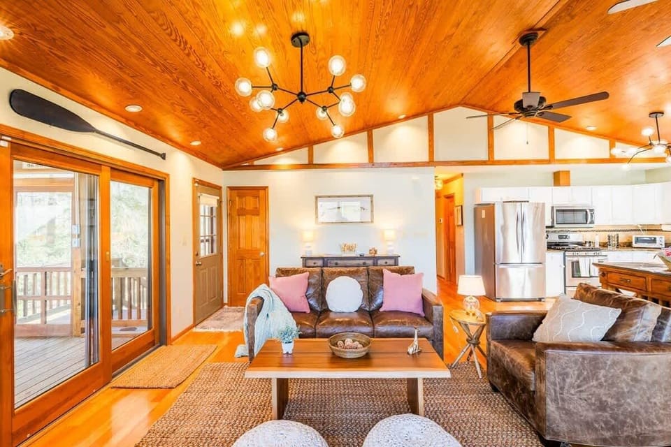 The waterfront living room with vaulted ceilings and a layout designed to maximize waterfront views combines the charm of a cabin retreat with the tranquility of waterfront living, providing a comfortable and memorable vacation experience. A modern chandelier with warm-toned bulbs complements the cabin aesthetic, while a woven rugs and wooden accents enhance the cabin feel.