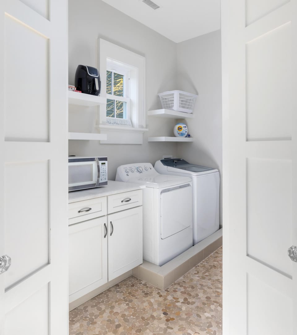 Laundry room with a washer, dryer, and white cabinets. Open shelves hold a basket and small appliances. The floor has a patterned tile.