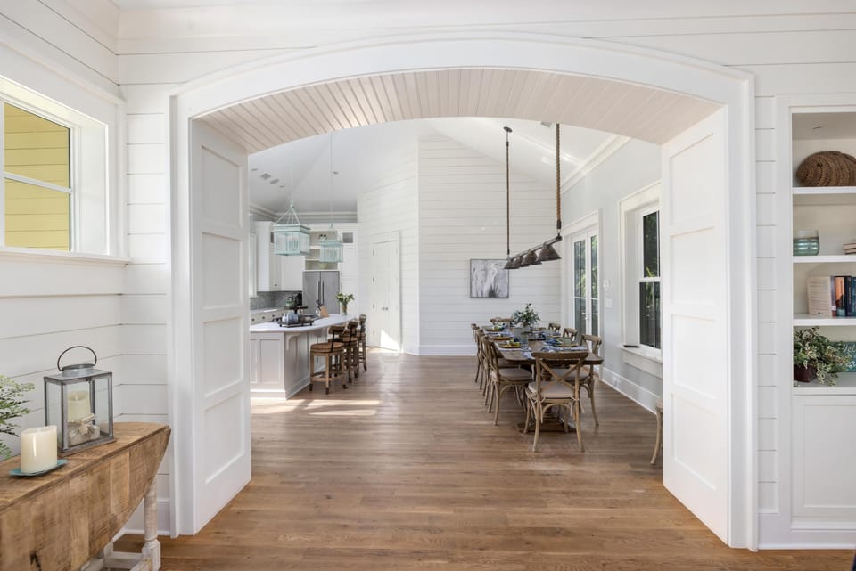 Bright open-concept kitchen and dining area with wooden floors, large table, pendant lights, white panel walls, and decorative shelves.