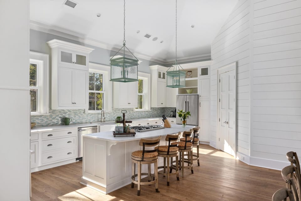 A spacious kitchen with white cabinets, a large island with wooden stools, pendant lights, and a view of trees through the windows.