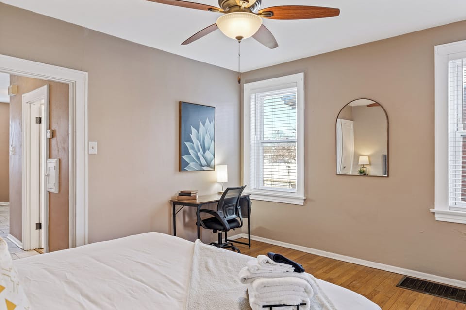 Bedroom view showing the edge of the bed, a desk with rolling chair beneath the window, two wall mirrors, hardwood flooring, and a ceiling fan.