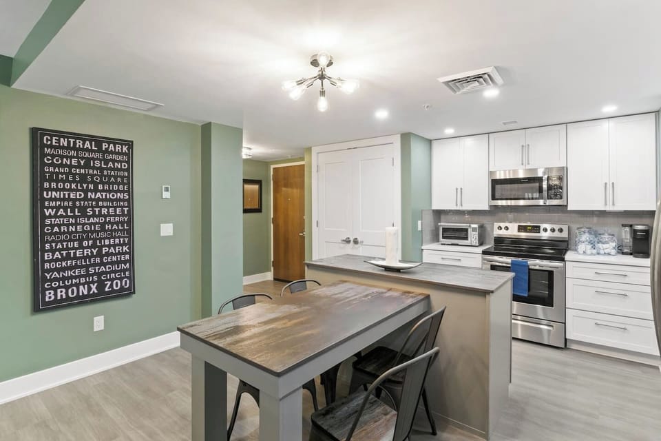 A modern kitchen with white cabinets, stainless steel appliances, and a dining area with a wooden table and chairs, accentuated by a statement wall listing iconic locations, under a bright ceiling light fixture.