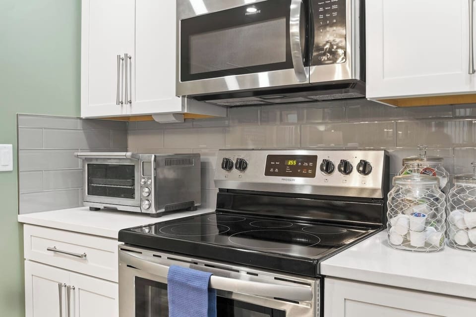 This is a well-equipped kitchen featuring a stainless steel stove and microwave, a toaster oven, and glass jars with coffee pods. The space is finished with white cabinets and a grey tile backsplash.