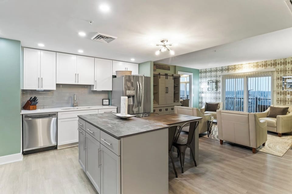 A spacious open-plan living area featuring a sleek kitchen with modern appliances and an island, a wooden dining table with chairs, leading to a cozy lounge area with comfortable seating and natural light streaming through large glass doors.