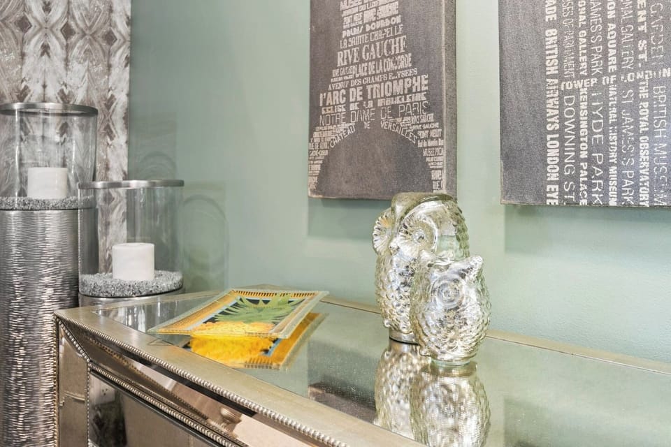 A mirrored console table that reflects its surroundings, enhancing the light and space of the room. City-themed wall art depicts famous locations in Paris and London, contributing to the cosmopolitan feel of the decor. The overall aesthetic is one of