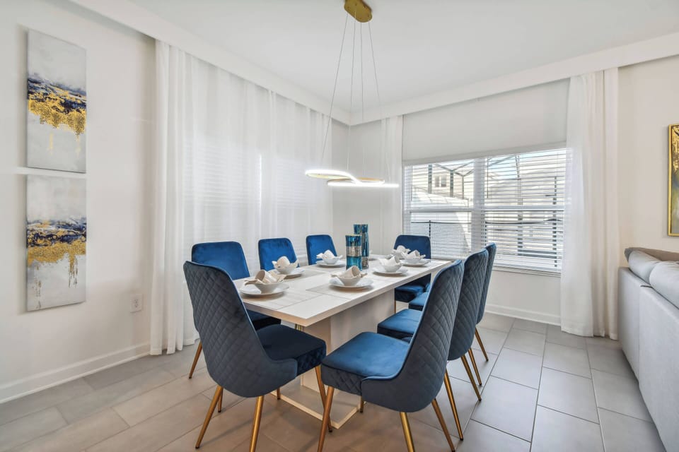 Gather around the stylish dining table set for six, featuring sleek blue chairs that pop against the light, airy backdrop. Soft natural light filters through large windows, enhancing the modern decor and inviting atmosphere.