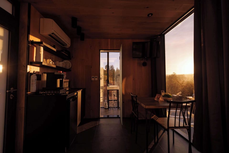 Warm evening light inside the Mirror House, with a cozy bed, dining area, and floor-to-ceiling windows surrounded by canyon scenery.
