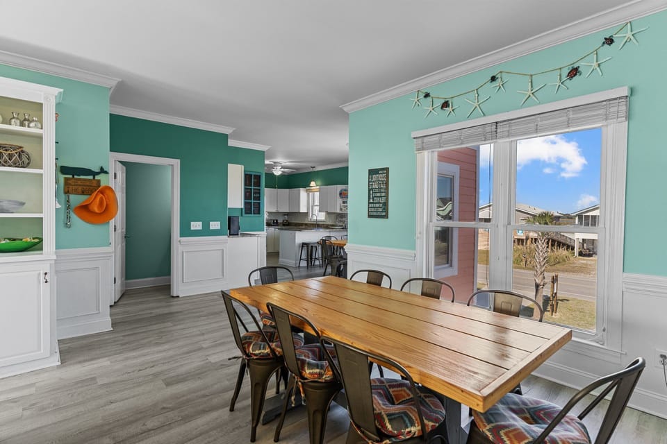Oak Island, NC dining room with large wood table, colorful cushioned chairs, teal walls, bright windows, beach décor, and open view into the coastal kitchen.