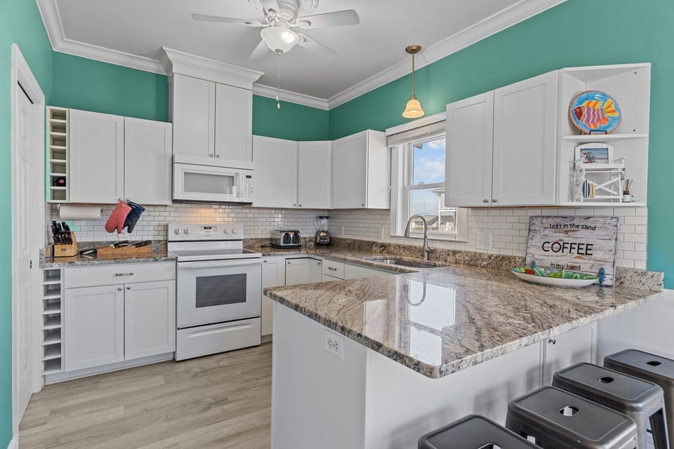 Oak Island, NC kitchen with teal walls, white cabinets, granite island, stove, microwave, sink, stools, coffee décor, subway tile, and beachy style.