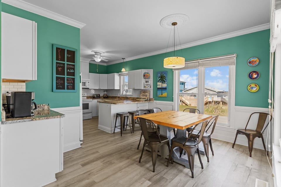 Oak Island, NC dining area with wood table, metal chairs, teal walls, large windows, pendant light, mirror, coastal plates, and sunny island street views.