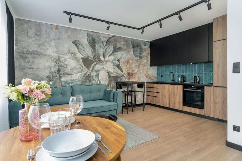 A wide shot of the kitchen and living area, featuring a floral mural, teal backsplash, and wooden cabinetry.

