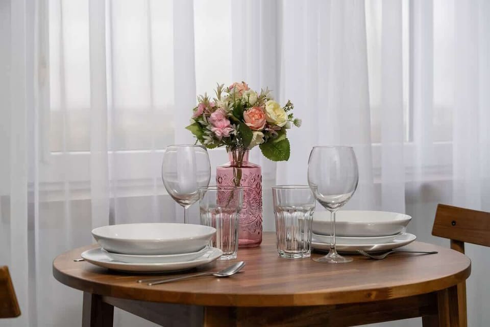 A close-up of a round dining table set with glasses, plates, and flowers near a window.

