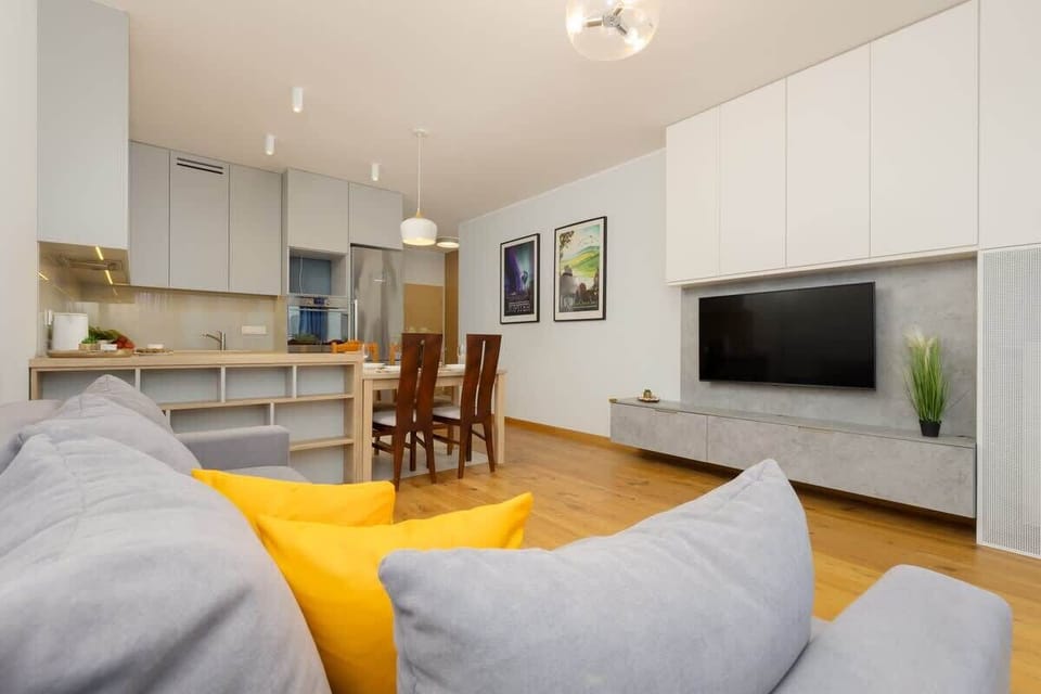An open-plan living and dining area with a grey sofa, yellow cushions, and a modern kitchen with white cabinets, maintaining a clean and functional design.
