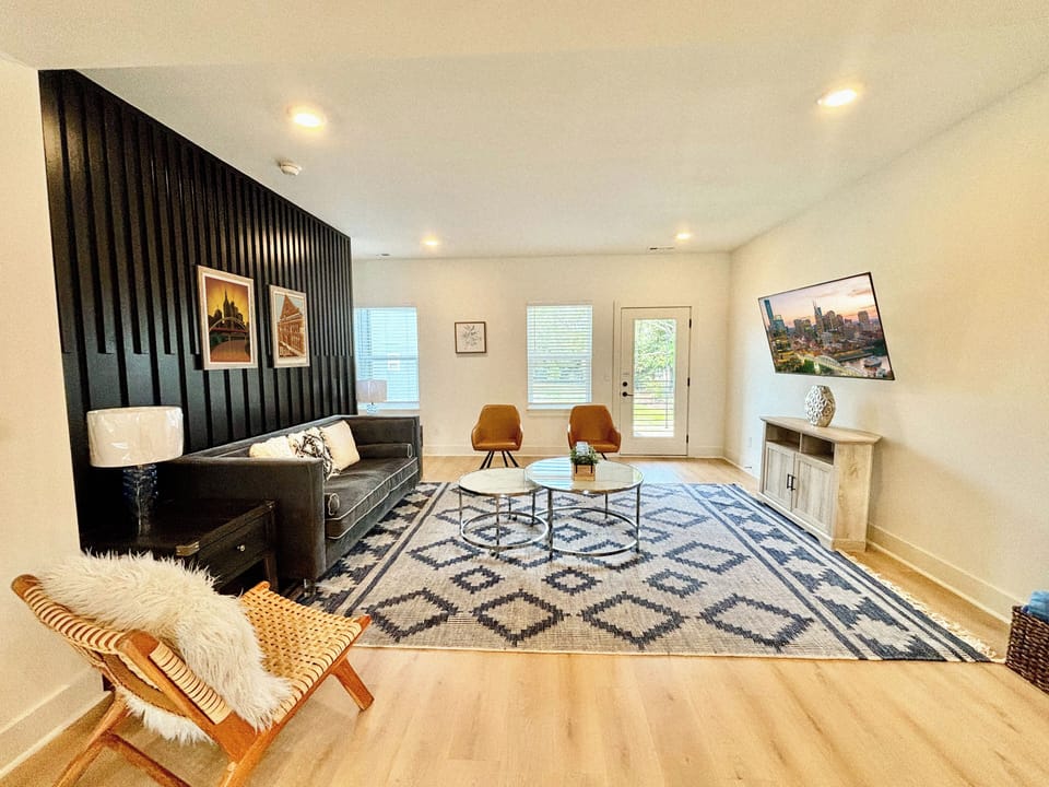 LIVING ROOM – A chic blend of modern and cozy, this living room is the perfect space for quiet relaxation or intimate gatherings. The sleek black accent wall draws the eye to the elegant velvet sofa, framed by soft lighting from the matching table lamps. Two mustard yellow chairs and a woven lounge chair offer stylish seating options, complemented by the geometric patterned are...