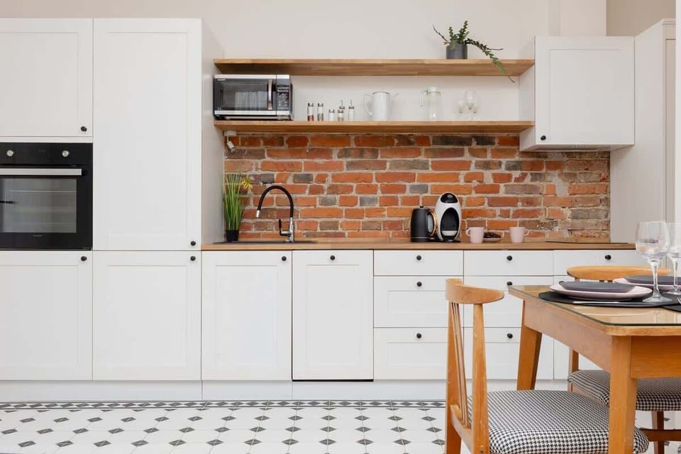 A minimalist kitchen with white cabinets, open wooden shelves, and a brick backsplash, blending modern and rustic elements.
