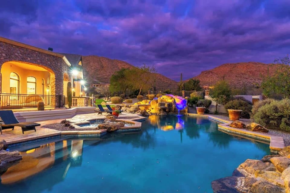 Evening settles over the stunning pool area, where a dramatic waterfall and rock grotto glow against desert mountain views and vibrant twilight skies.