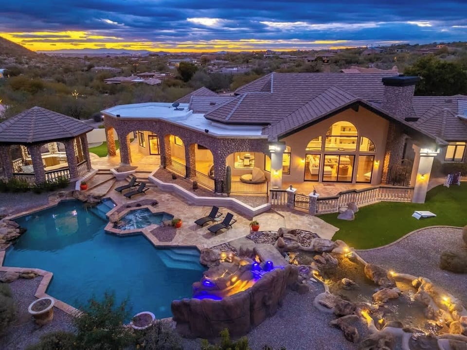Spectacular desert estate at twilight, featuring an expansive stone villa with a custom pool, grotto-style hot tub, and waterfall features set against a dramatic Arizona sunset and distant mountain views.