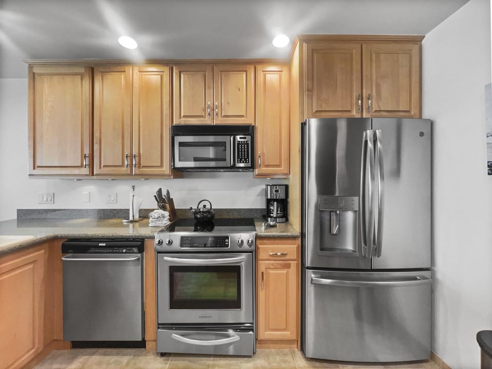 A modern kitchen with stainless steel appliances, including a refrigerator, oven, microwave, dishwasher, and coffee maker. Light wood cabinets and a granite countertop complete the setup.