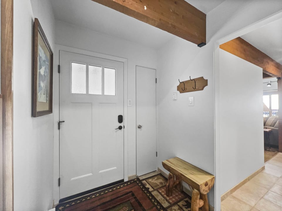 A small entryway features a white door with frosted glass panels, a wooden bench, a wall-mounted coat rack, a colorful rug, a piece of framed art, and a view into a living area.