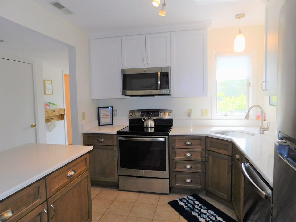 543C hudspn Ave, Stewards Watch, Bethany Beach - The kitchen has been fully modernized with a two-tone cabinetry style—clean white uppers and warm wood-toned lowers—paired with stainless steel appliances and white quartz countertops. The open-concept layout includes a breakfast bar that looks through to the dining area, creating a social flow that is highly desirable for coastal living.