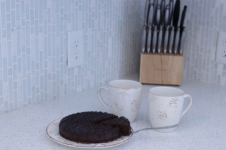 Quartz countertop with two shell-accent mugs, chocolate cake, and knife set against tiled backsplash—perfect cozy coffee setup in this bright Oak Island beach kitchen.