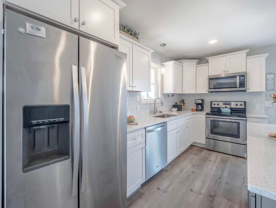 Bright kitchen with white cabinetry, stainless steel appliances, large island with seating, pendant lighting, and door to the deck—perfect for cooking and gathering in this Oak Island beach home.