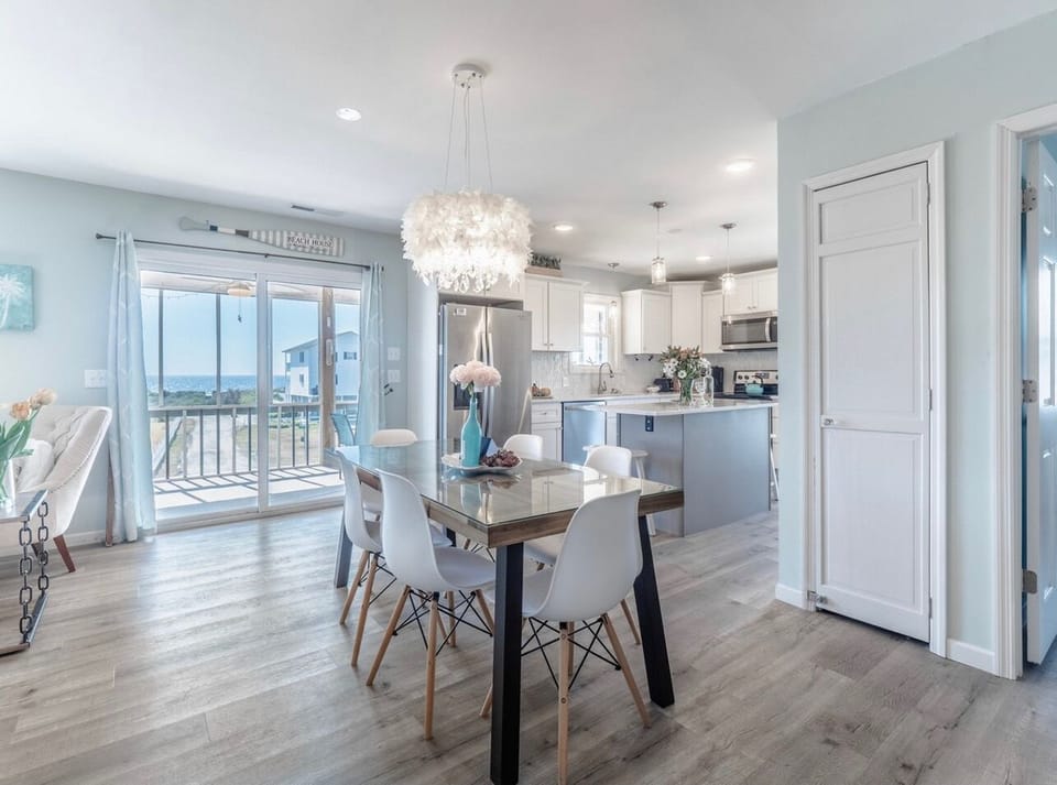 Bright dining area with modern table, stylish lighting, and open kitchen with stainless appliances, plus sliding doors to a deck with ocean views—perfect for meals at this Oak Island beach home.