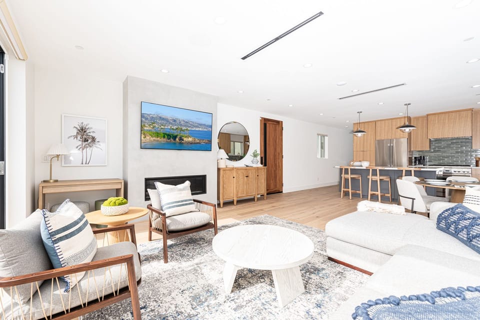 The living room features plenty of seating, a TV, and an open layout connecting it to the kitchen and front patio.
                