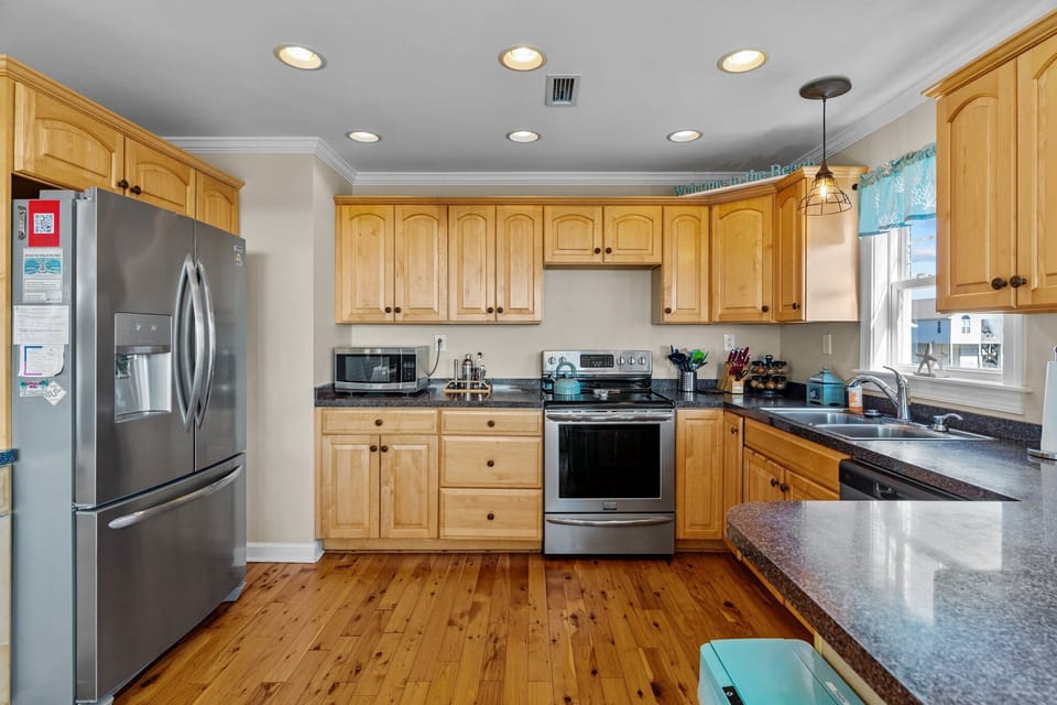 Oak Island, NC beach rental kitchen with light wood cabinets, stainless refrigerator and stove, microwave, dark countertops, double sink under window, pendant and recessed lighting, hardwood floors, knife block, coffee items, and coastal decor.