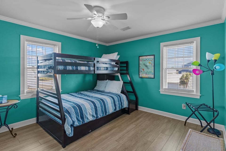 Bright Oak Island, NC bunk bedroom featuring a twin-over-full bunk bed, teal walls, coastal bedding, ceiling fan, wood-look floors, and two sunny windows. Fun, comfortable space ideal for kids or extra guests.