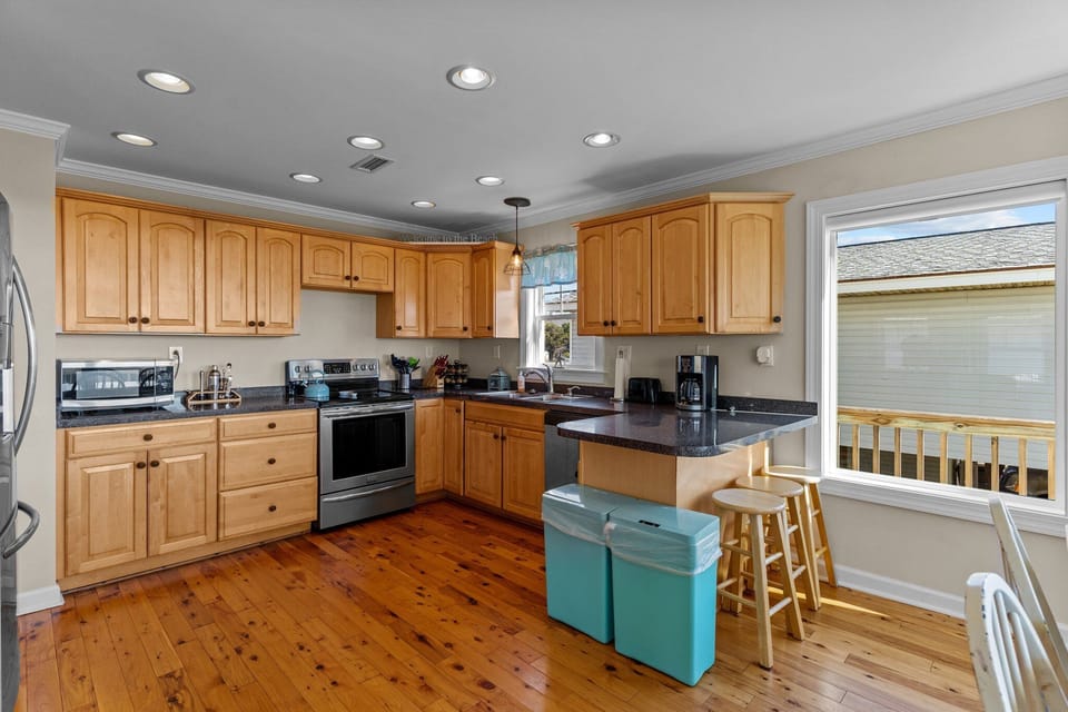 Oak Island, NC vacation rental kitchen with light wood cabinets, stainless stove and fridge, dark counters, island breakfast bar with stools, coffee maker, microwave, sink under window, recessed lighting, hardwood floors, and deck view outside.