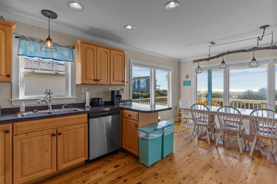 Oak Island, NC beach rental kitchen with light wood cabinets, stainless refrigerator and stove, microwave, dark countertops, double sink under window, pendant and recessed lighting, hardwood floors, knife block, coffee items, and coastal decor.