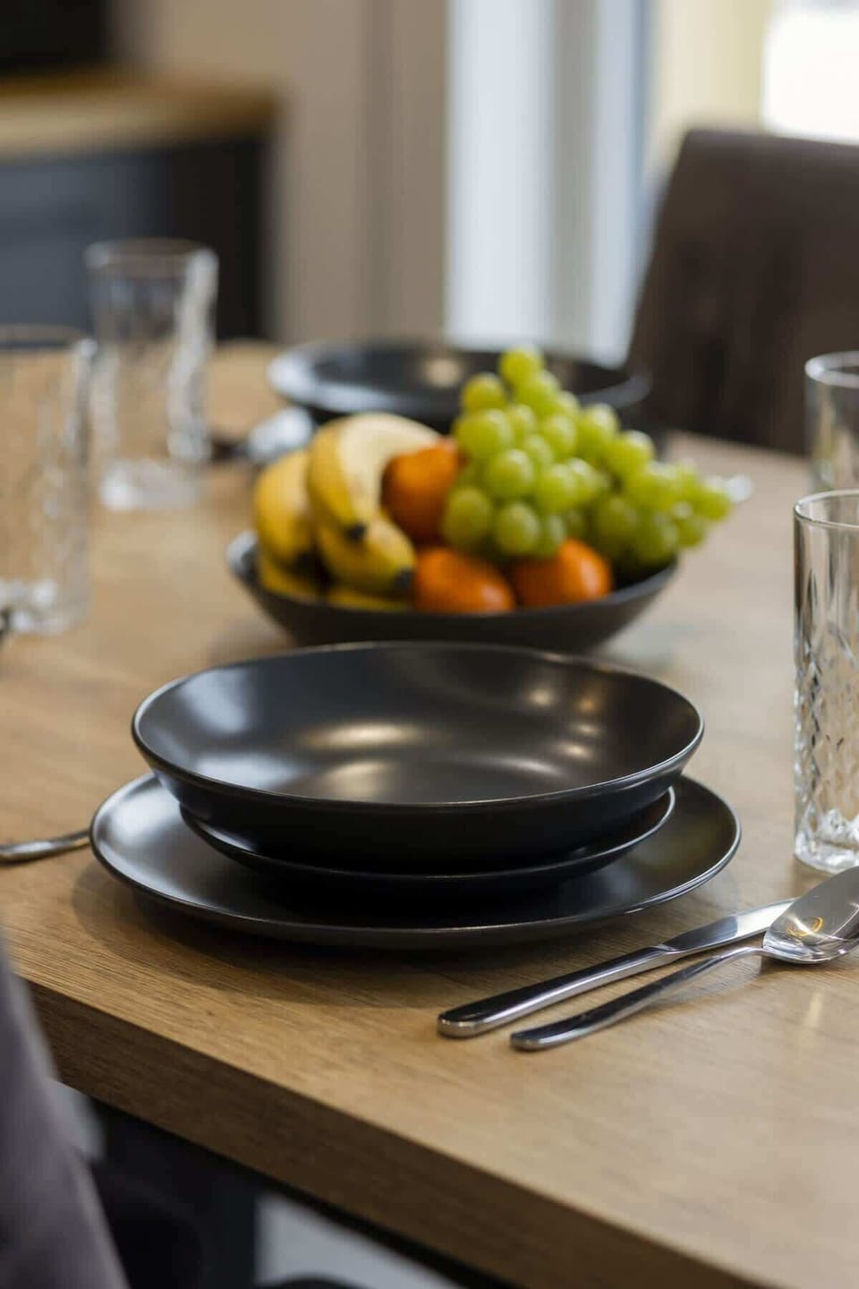Beautifully set dining table with black plates, gold cutlery, and fresh fruit centerpiece.
