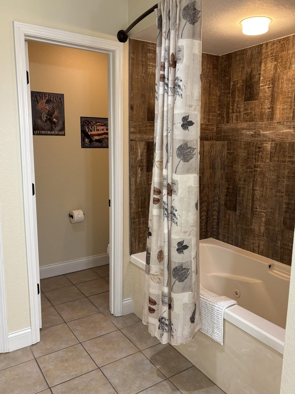 Bathroom with tiled floor, jetted bathtub surrounded by wood-look panels, and leaf-patterned shower curtain.