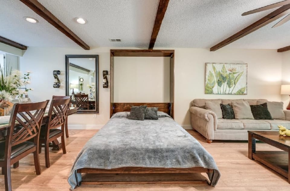 Murphy bed with gray blanket folds into wood frame beside beige sofa, dark wood coffee table, and dining chairs.