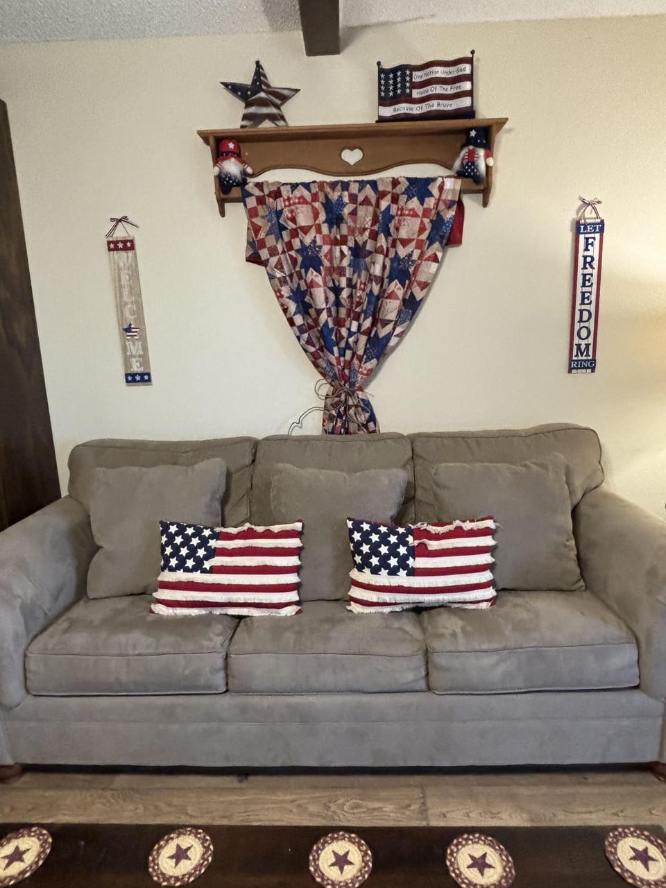 A gray fabric sofa is accented with two American flag pillows beneath a wooden wall shelf displaying patriotic decorations.