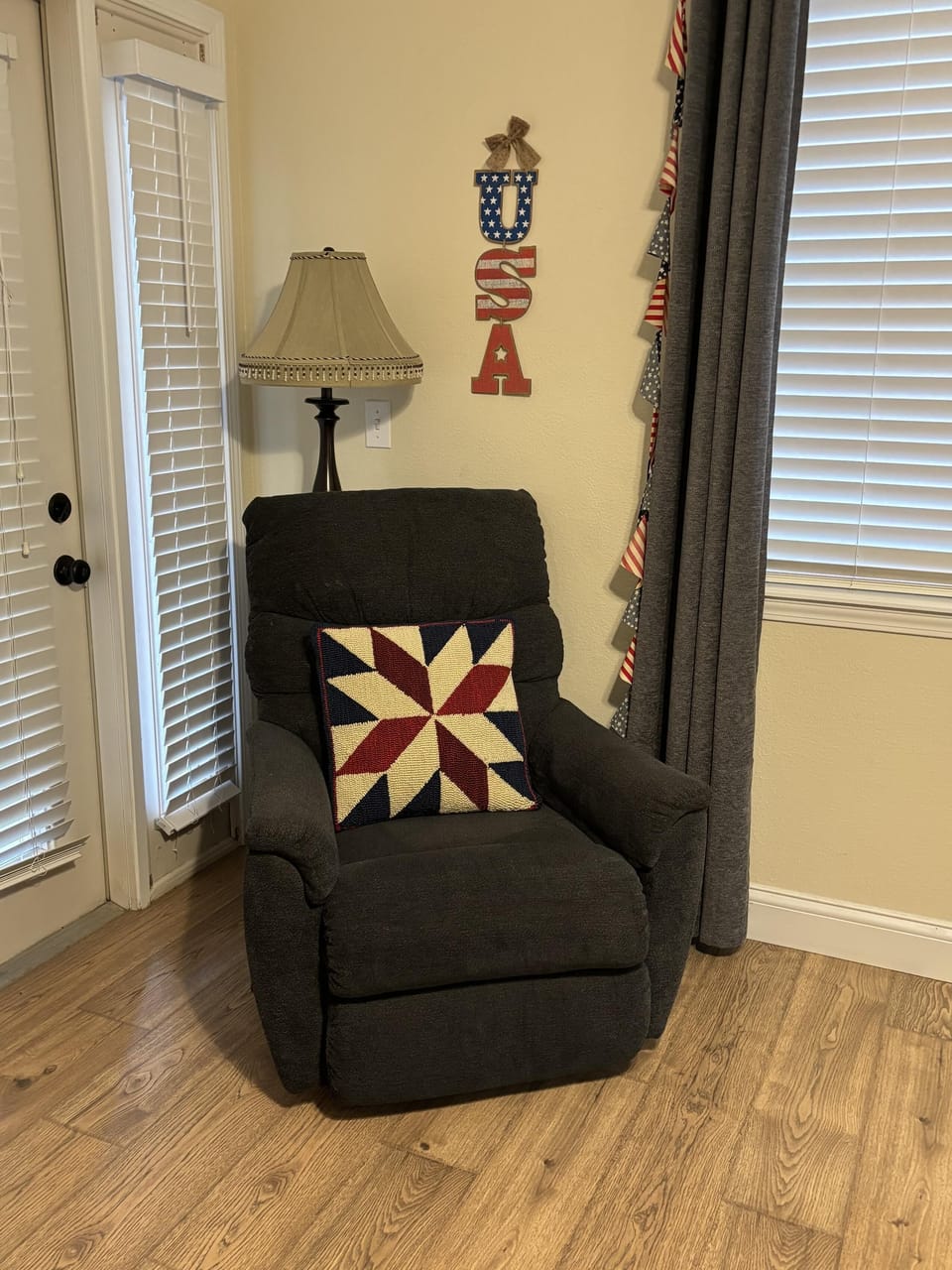 Dark gray recliner with star-patterned cushion sits beside a floor lamp near French doors and patriotic wall decor.