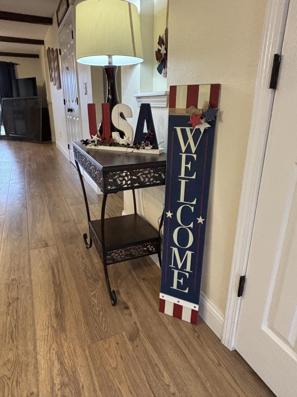 Iron console table with decorative cutouts holds a lamp and USA letters, next to a tall red, white, and blue welcome sign.