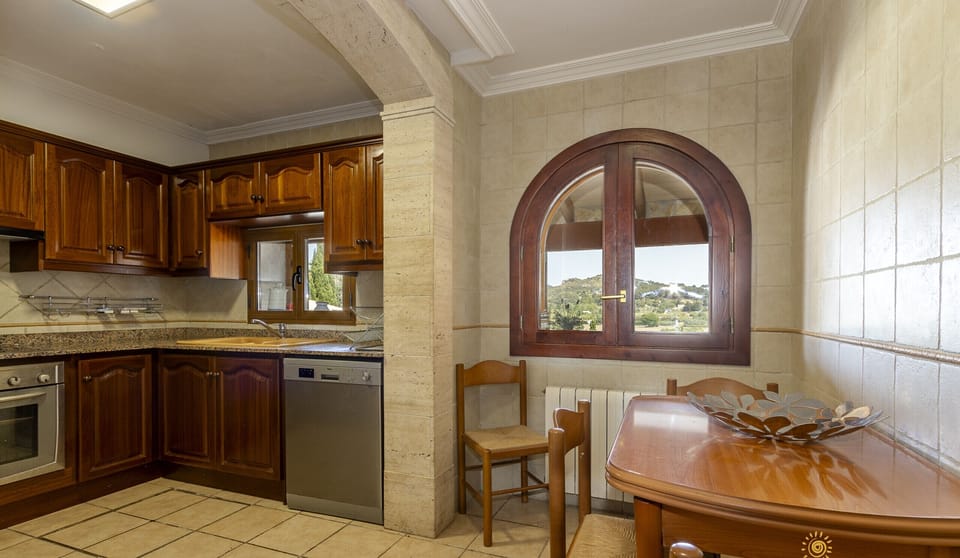 Kitchen at Villa Alejandra with integrated dining area, wooden cabinets, dining table, fully equipped appliances and a window with views of the natural surroundings.