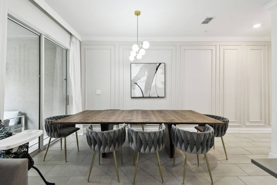 Come together at the beautiful wooden dining table, surrounded by cozy, textured chairs perfect for enjoyable conversations. Large windows let in bright light, enhancing the elegant wall panels and the modern chandelier above.