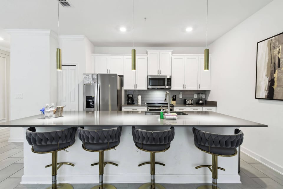 Welcome to a stylish kitchen featuring sleek gray countertops and white cabinetry, where modern appliances and dark-fabric bar stools create comfort, all bathed in warm pendant lighting for delightful moments with loved ones.