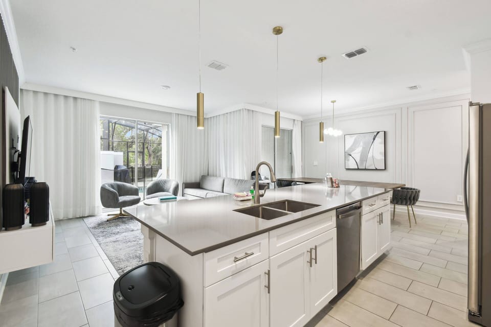 Enjoy a bright kitchen with sleek white cabinetry and stainless steel appliances that leads to a cozy living area filled with natural light from large windows, complemented by stylish pendant lighting for a warm, inviting.