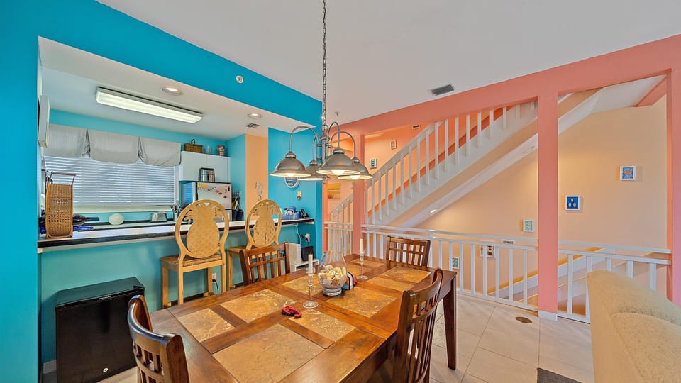 A brightly colored dining area with a wooden table and six chairs next to an open kitchen. The space features vibrant blue and coral walls, white stairs, and modern pendant lighting.