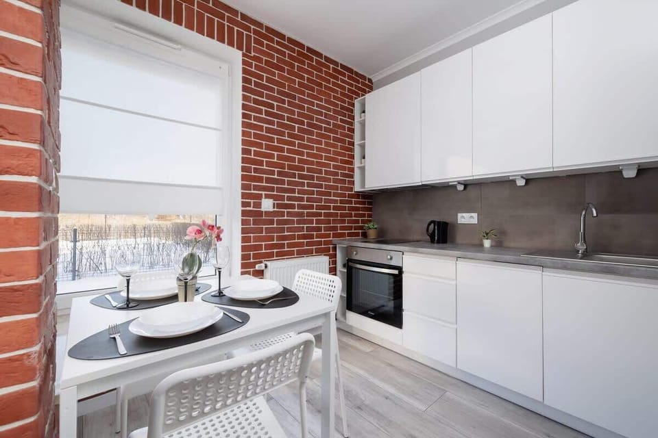 A contemporary kitchen and dining area with sleek white cabinets, a compact dining table, and an exposed brick wall adding a rustic charm.
