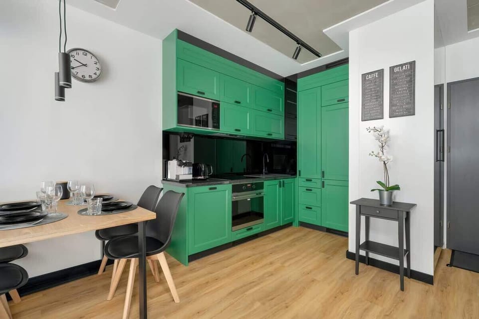 An open-plan setup showcasing a striking green kitchen, a small dining table, and gray walls that complement the wooden floor.
