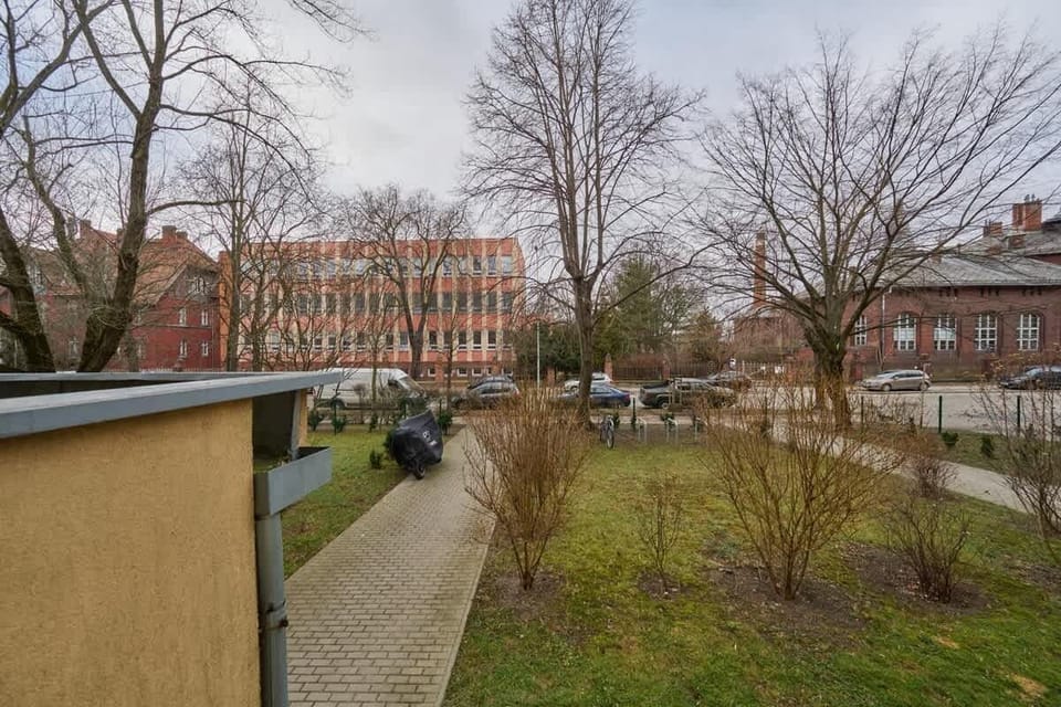 An outdoor walkway with greenery and trees, leading to a residential area.
