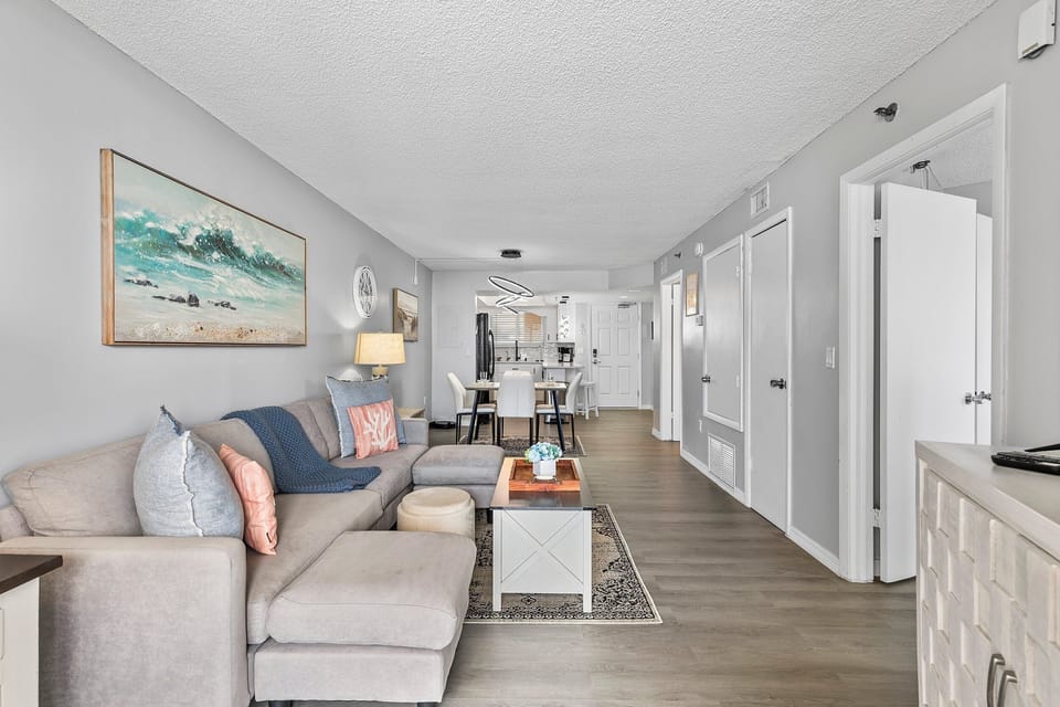 A plush sectional sofa with coral and ocean-blue accents provides comfort, while the large beach-themed artwork adds a serene, seaside touch.